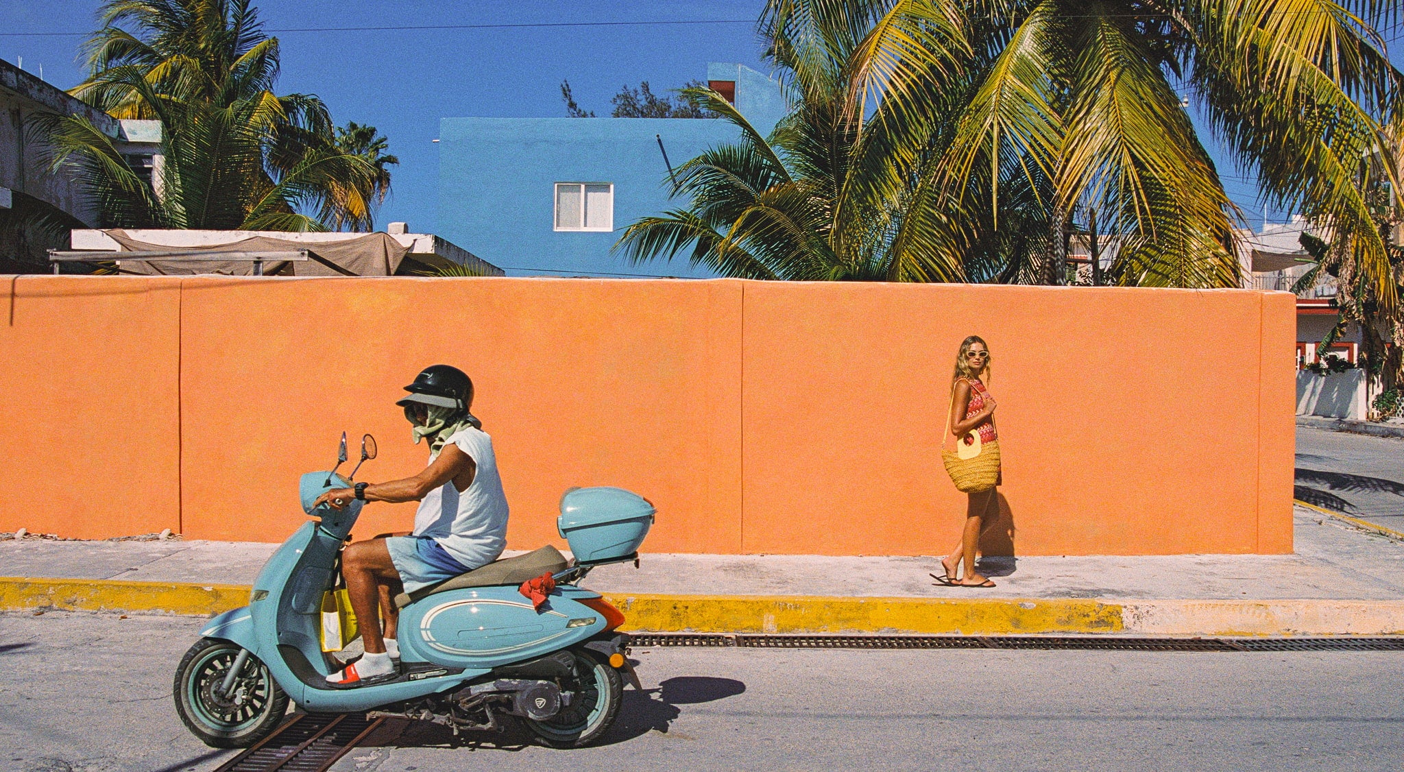 Person on a blue scooter in front of an orange wall with a woman standing nearby.