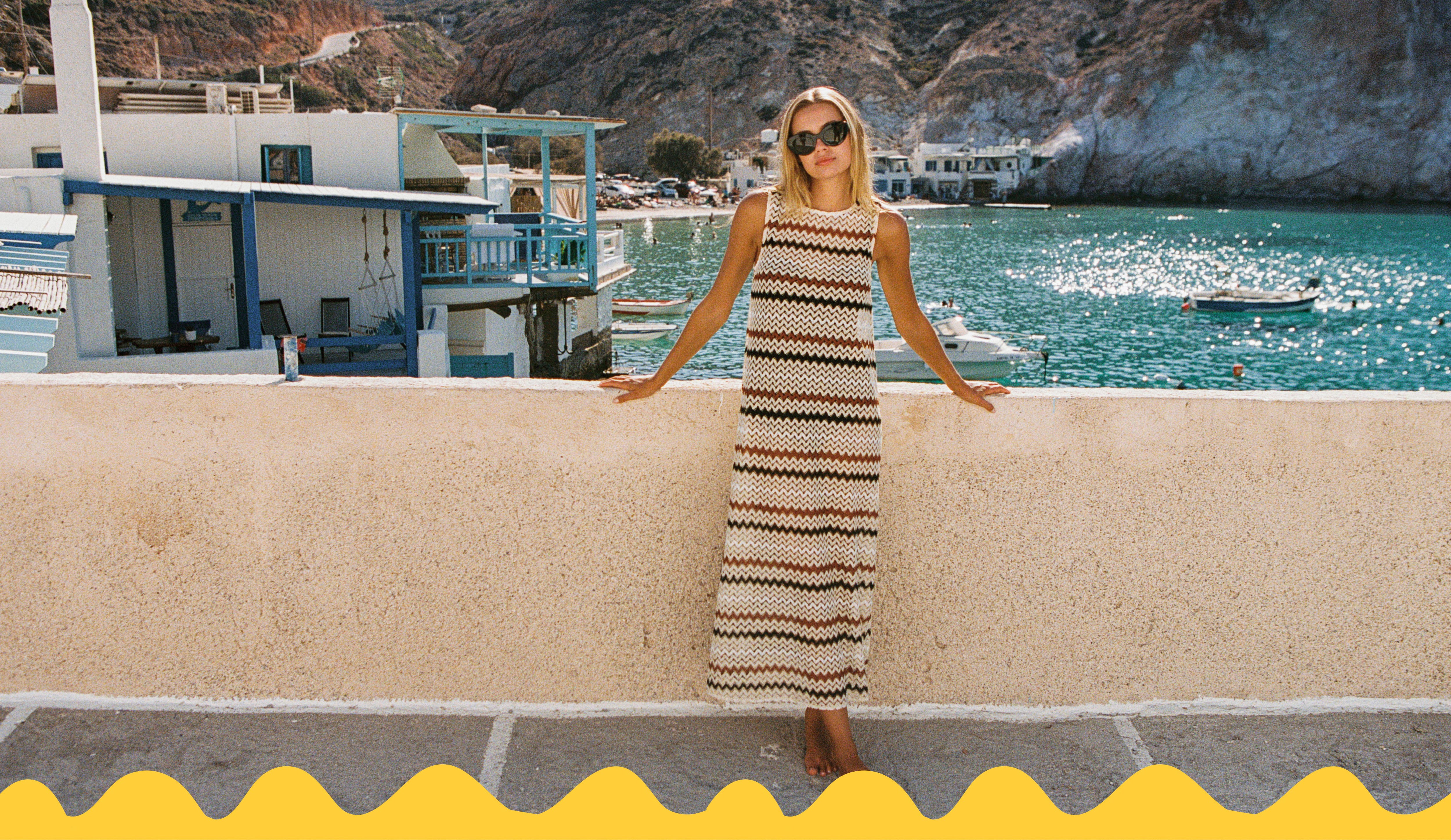 A woman wearing a sleeveless beige and black patterned maxi dress and sunglasses stands on a balcony overlooking a scenic coastal harbor with blue water, boats, and white buildings against rocky cliffs.