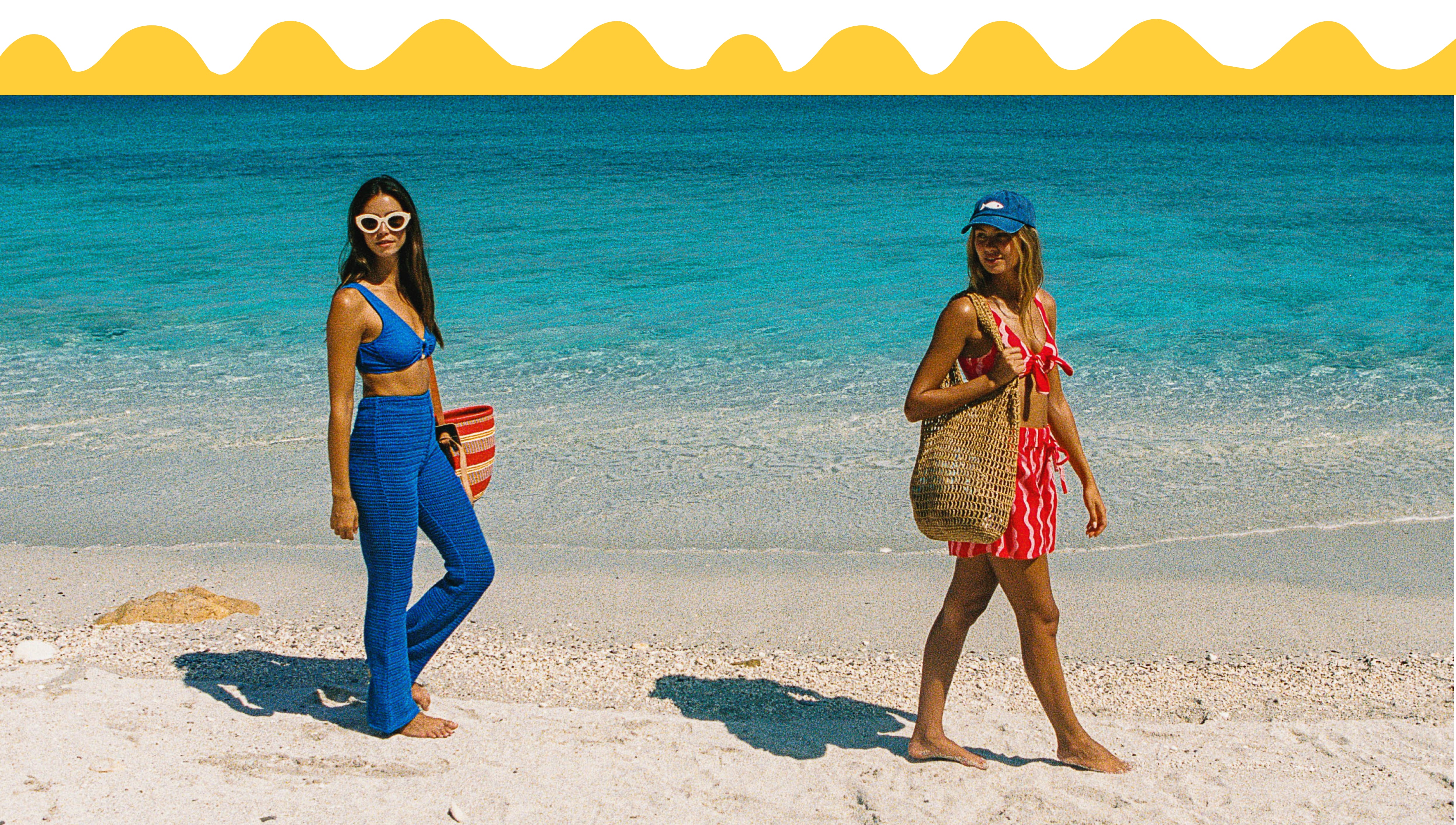 Two women walking on a sandy beach with turquoise ocean water in the background. One wears a bright blue two-piece swimsuit and holds a striped bucket bag, while the other wears a red and white striped bikini, a blue cap, and carries a woven tote bag.