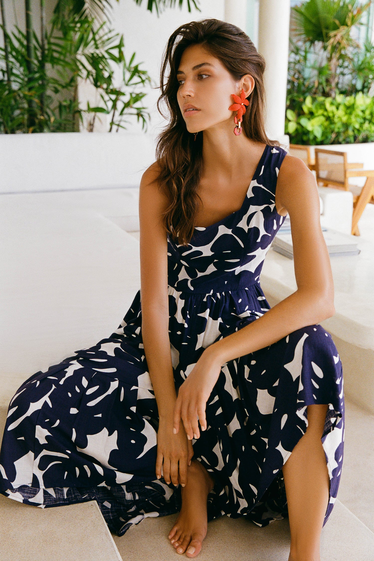 A woman wearing a navy and white tropical leaf print maxi dress sits on a white outdoor sofa. She is wearing bright orange flower earrings and looking off-camera.