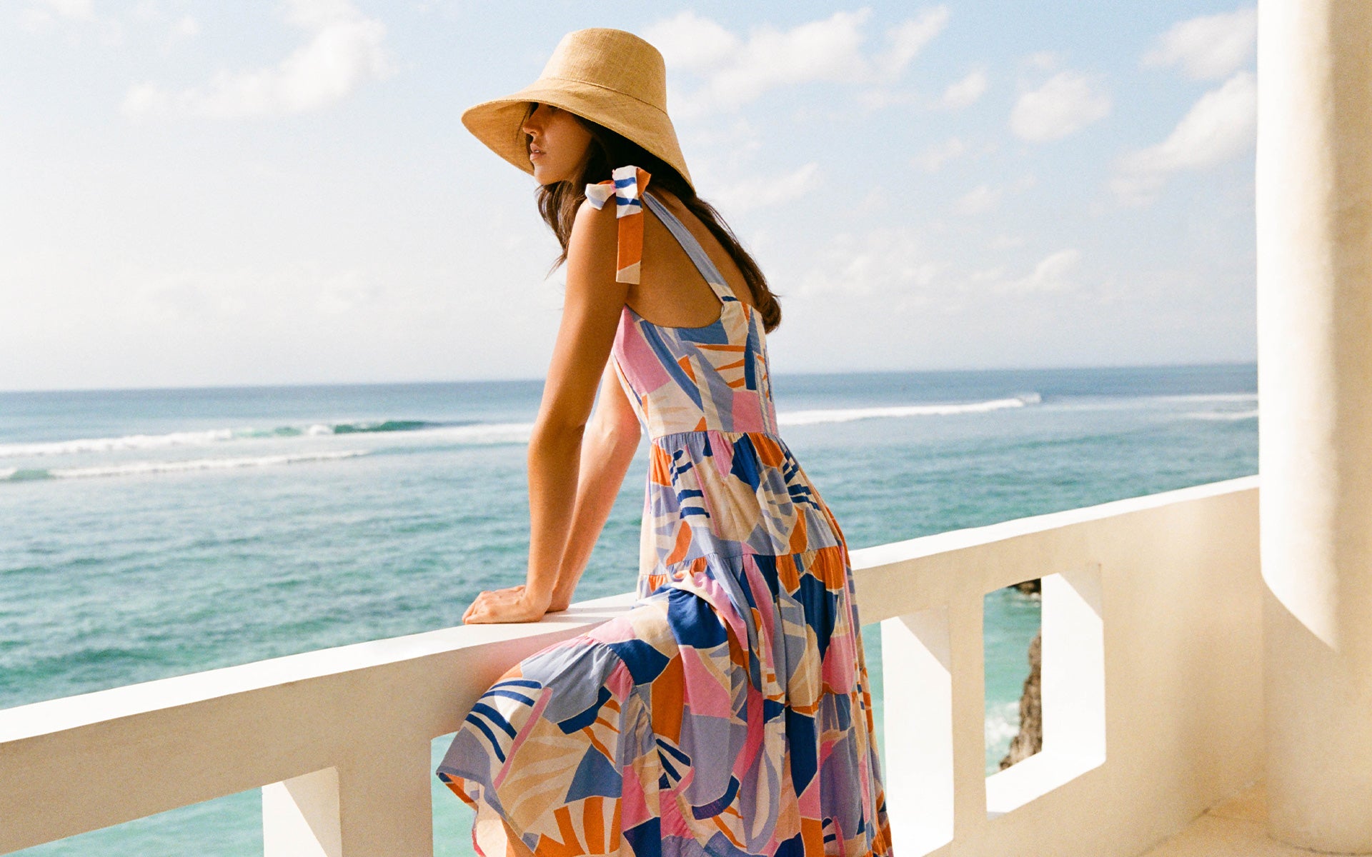 A woman wearing a straw hat and a colorful geometric print maxi dress leans on a white balcony railing with the ocean and sky in the background.
