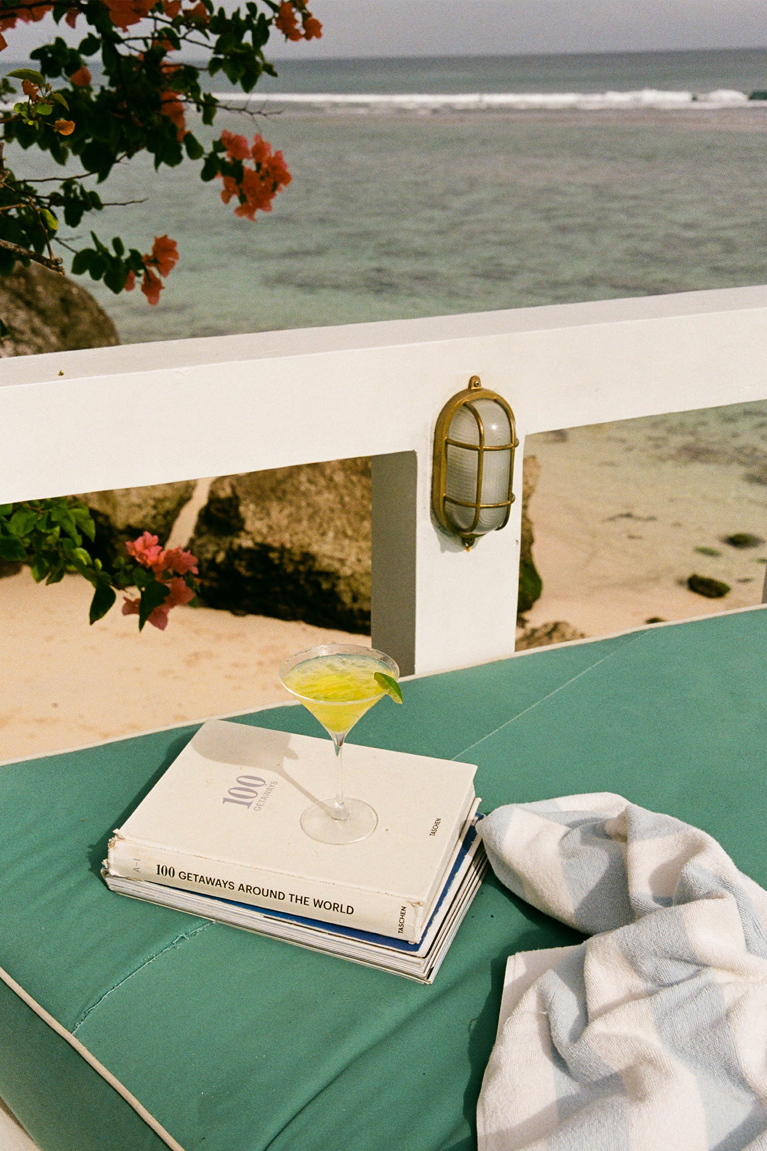 A styled beach lounge scene with a green cushion, a white railing, and the ocean in the background. On the cushion sits a stack of books with a yellow cocktail in a martini glass on top and a white towel nearby.