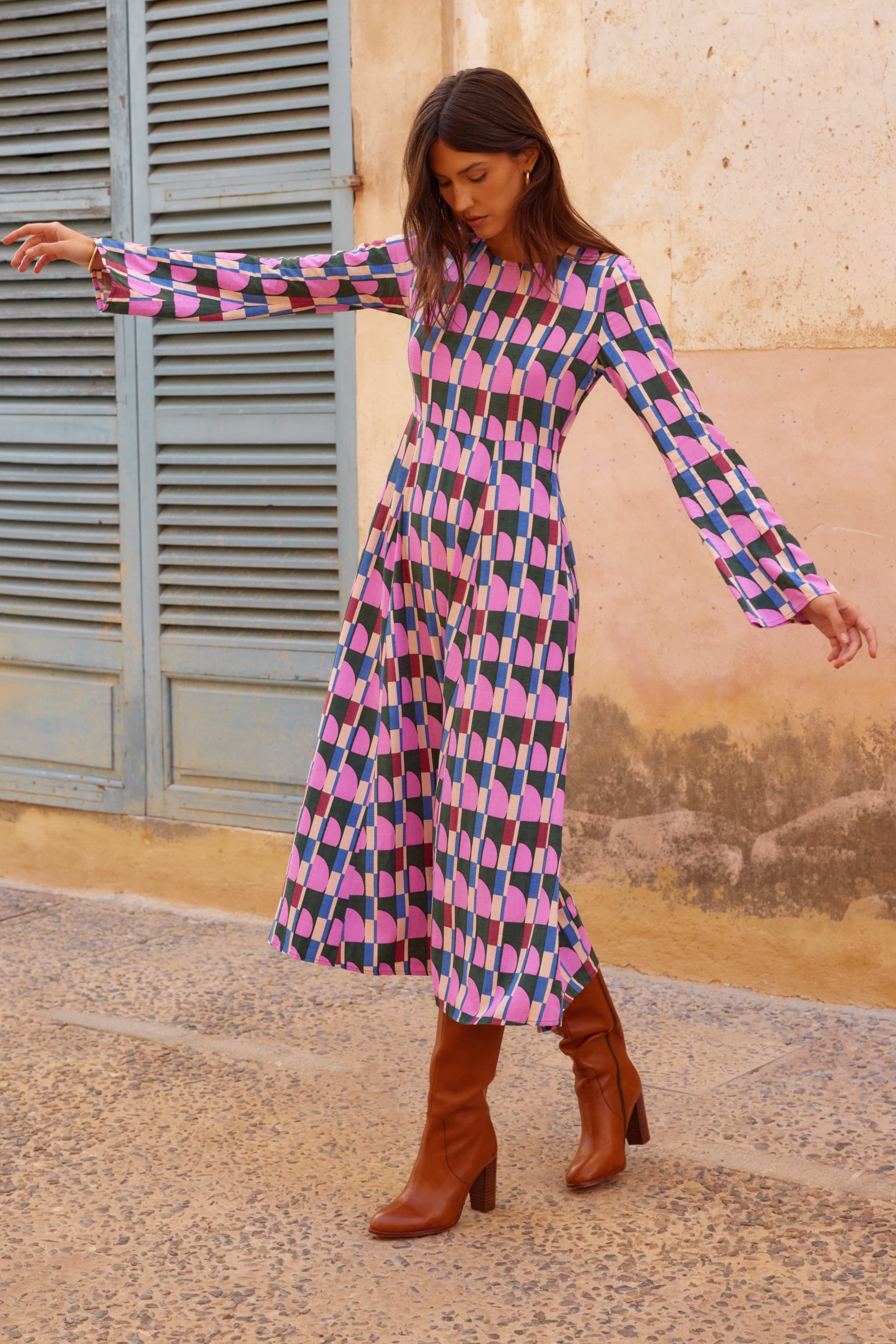 Woman wearing a colorful geometric dress with brown boots standing against a textured wall.