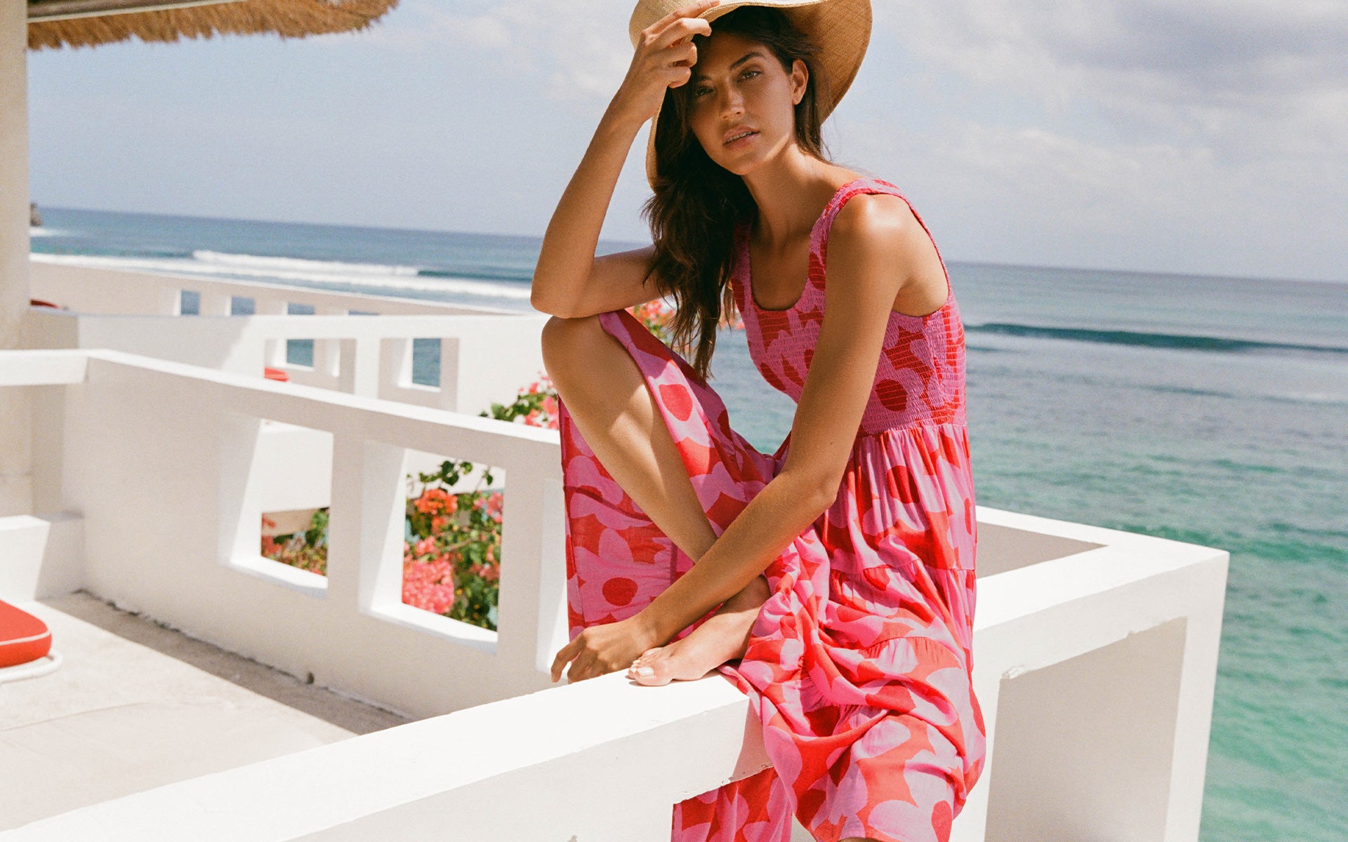 A woman wearing a pink and coral patterned sleeveless dress and a wide-brim straw hat sits on a white balcony overlooking a tropical ocean.