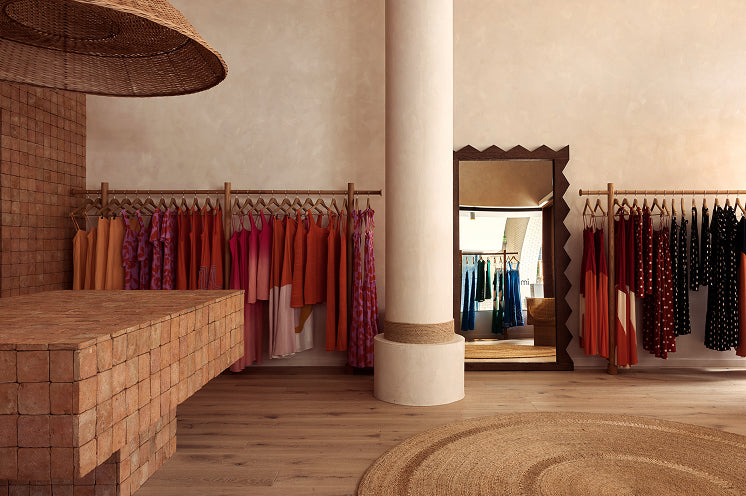 A boutique interior with clothing racks displaying colorful garments, a large cream column, a woven pendant light, and a round jute rug on a wood floor.