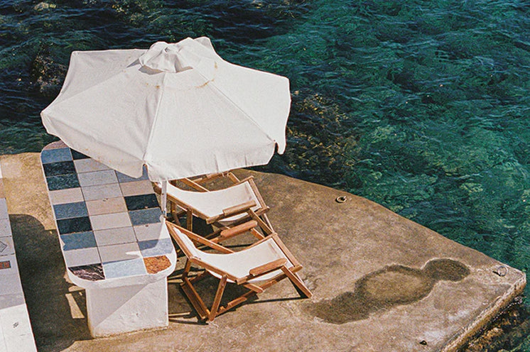 Outdoor furniture set with a white umbrella and chairs on a dock by the water.
