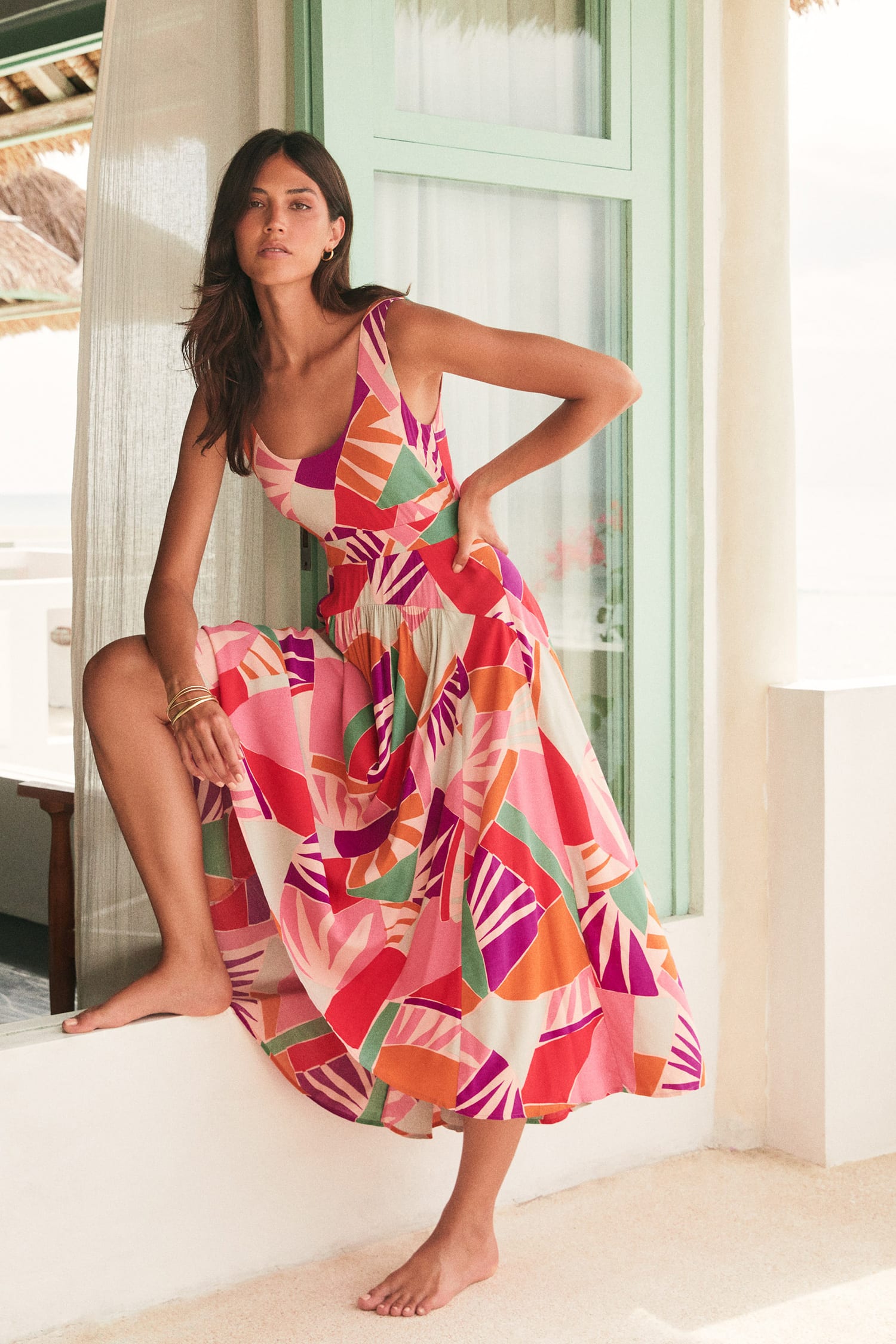 A woman models a sleeveless, flowy midi dress with a bold tropical leaf print in pink, orange, purple, green, and white, posing on a white balcony with a light green window in the background.