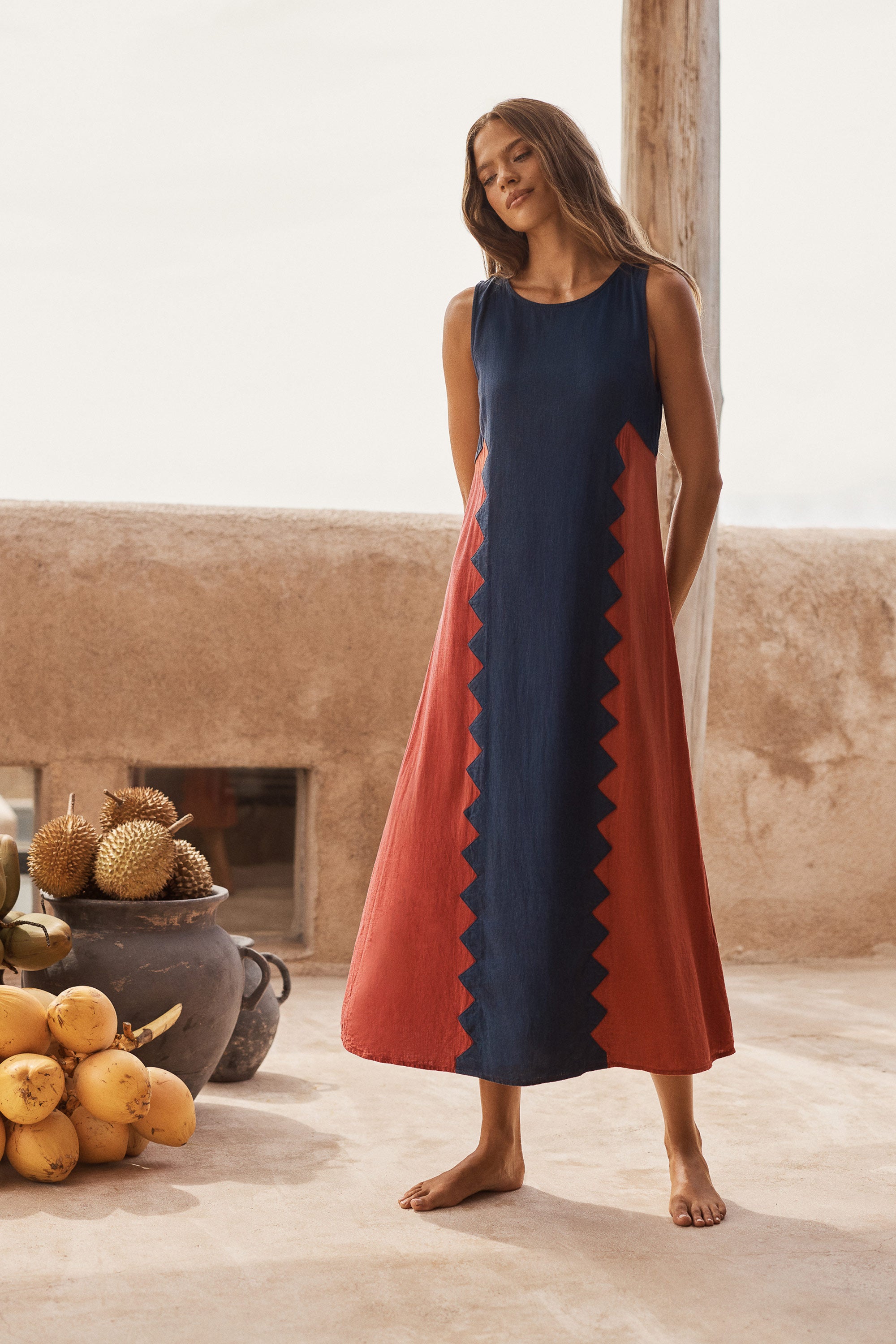 A woman stands barefoot in a rustic indoor setting wearing a sleeveless midi dress with a navy blue body and rust-red side panels separated by a zigzag border. The background features a textured beige wall, a wooden pillar, and a rustic clay pot with fruit.